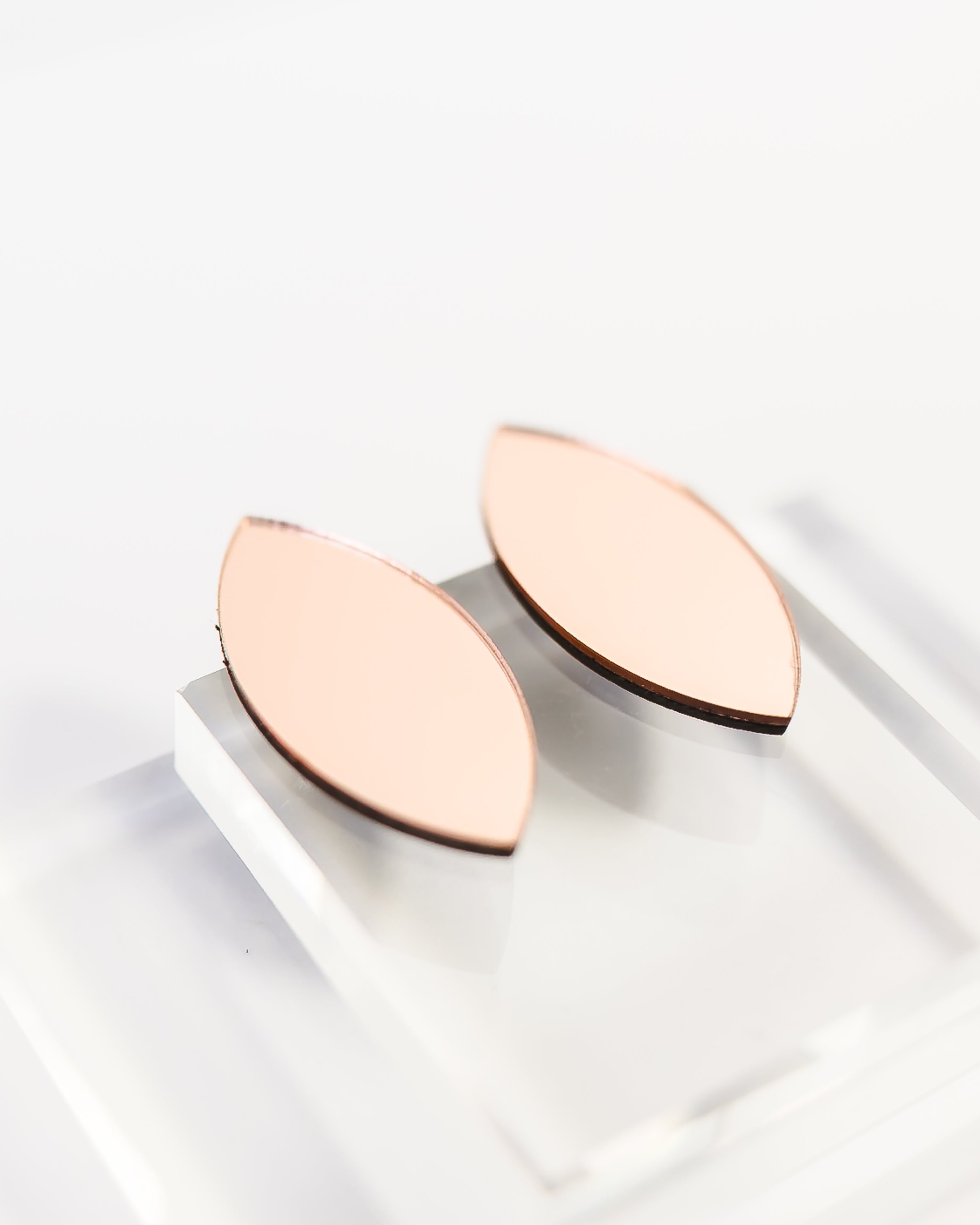 A pair of minimalist rose gold color mirror acrylic leaf-shaped earrings displayed on a clear acrylic riser against a clean white background.