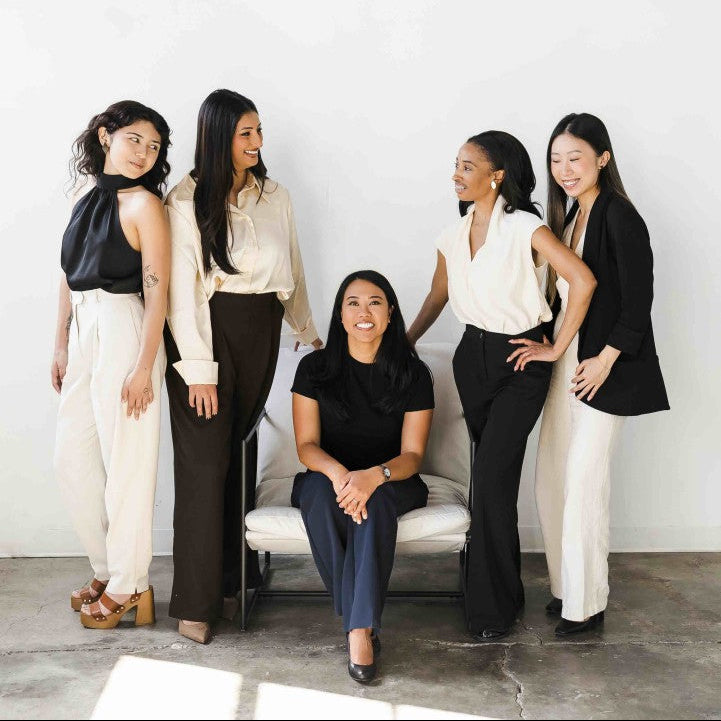 Four smiling women standing around one smiling woman who is sitting and facing the camera.