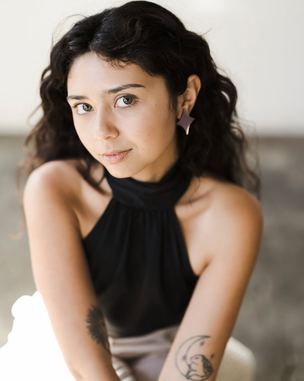 Young woman with long, curly dark hair sits facing camera, wearing black halter top and rose gold mirror earrings shaped like stars. Tattoos on arm.