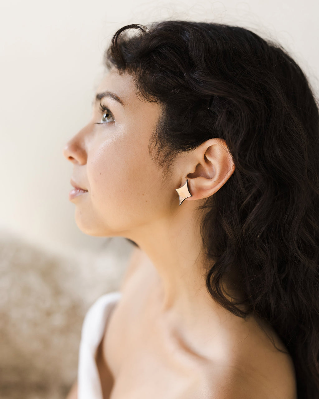 Woman with long, dark curly hair wearing a strapless white top. She wears a dainty satin gold acrylic earring and is gazing upward.