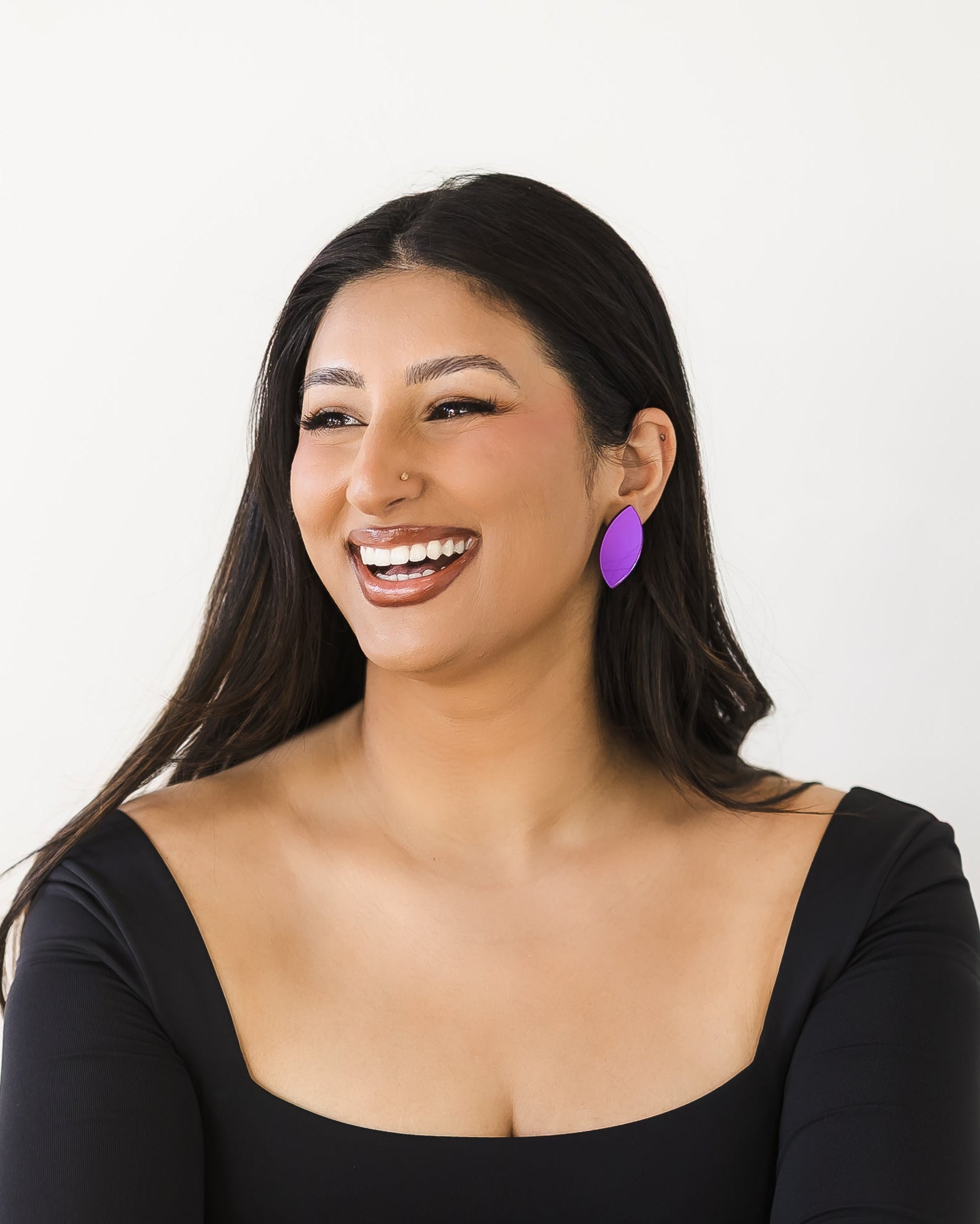 Woman with long dark hair wearing a black long-sleeve top and purple mirror acrylic leaf-shaped earrings, with a big smile.