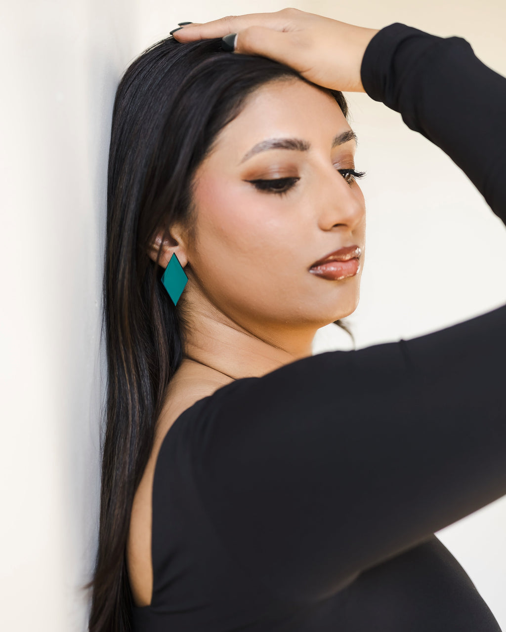 A woman with long black hair leans against a white wall, wearing reflective  bright teal geometric dangle earring.