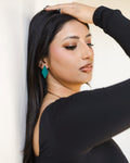 A woman with long black hair leans against a white wall, wearing reflective  bright teal geometric dangle earring.