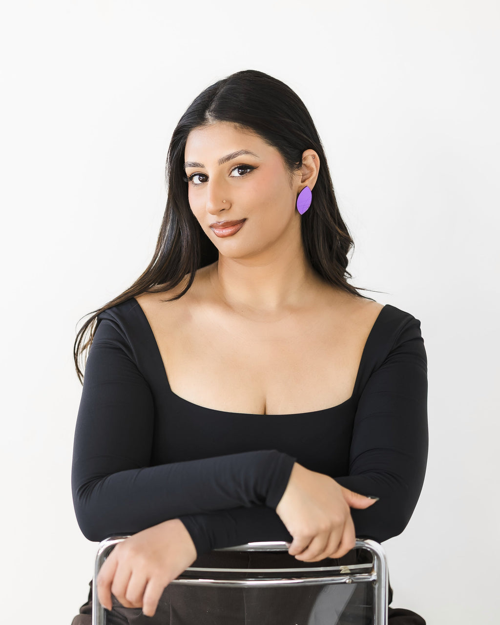Woman with long dark hair wearing a black long-sleeve top and purple mirror acrylic earrings, smiling demurely at camera.