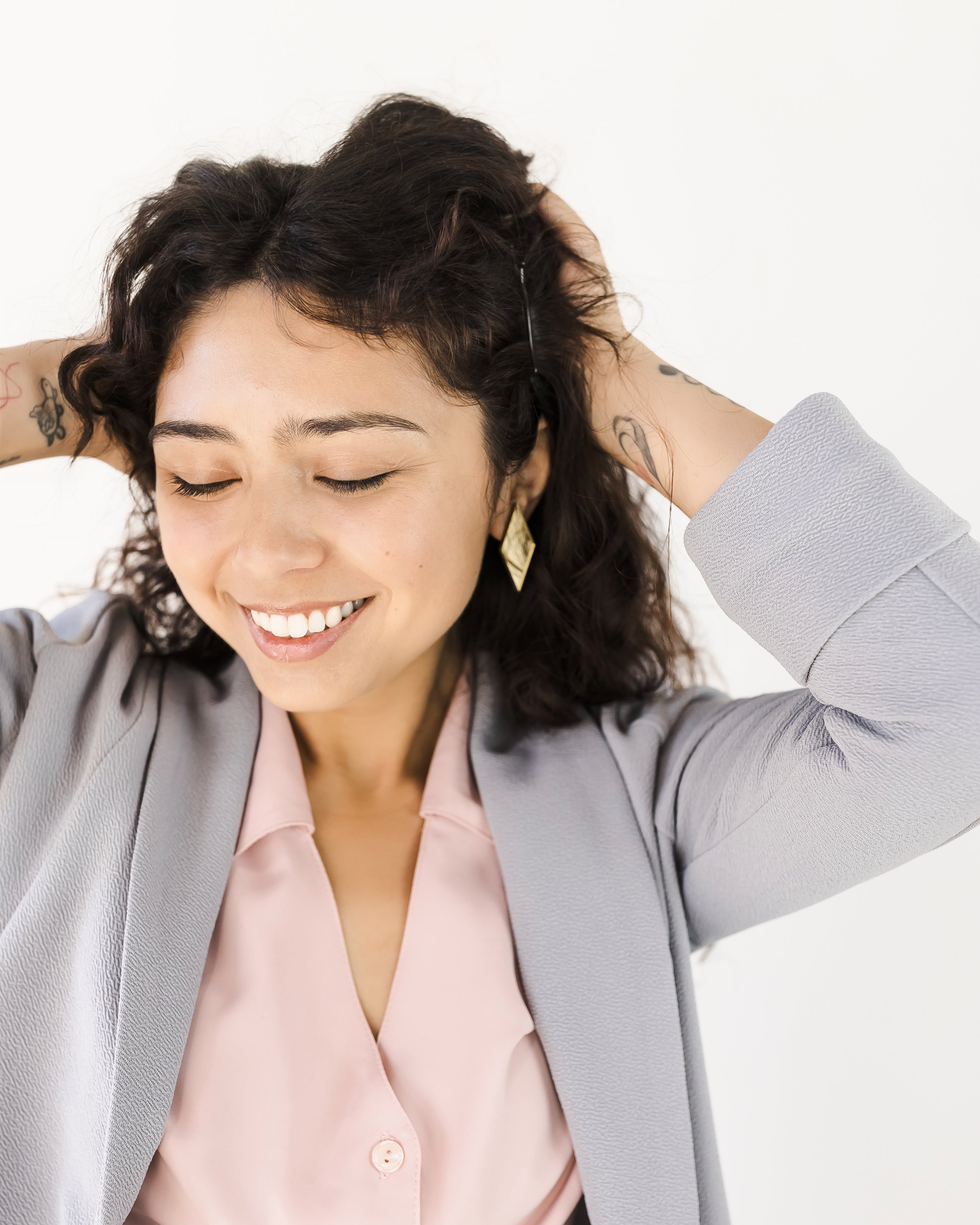 Woman with curly dark hair wearing a light gray blazer over a pale pink button-up blouse, posing with her hands in her hair against a plain white background.