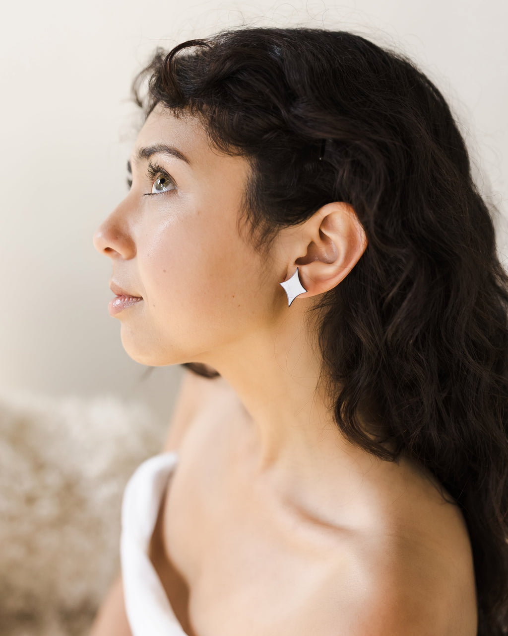 Woman with long, dark curly hair wearing a strapless white top. She wears a dainty sparkle earring and is gazing upward against a light background.