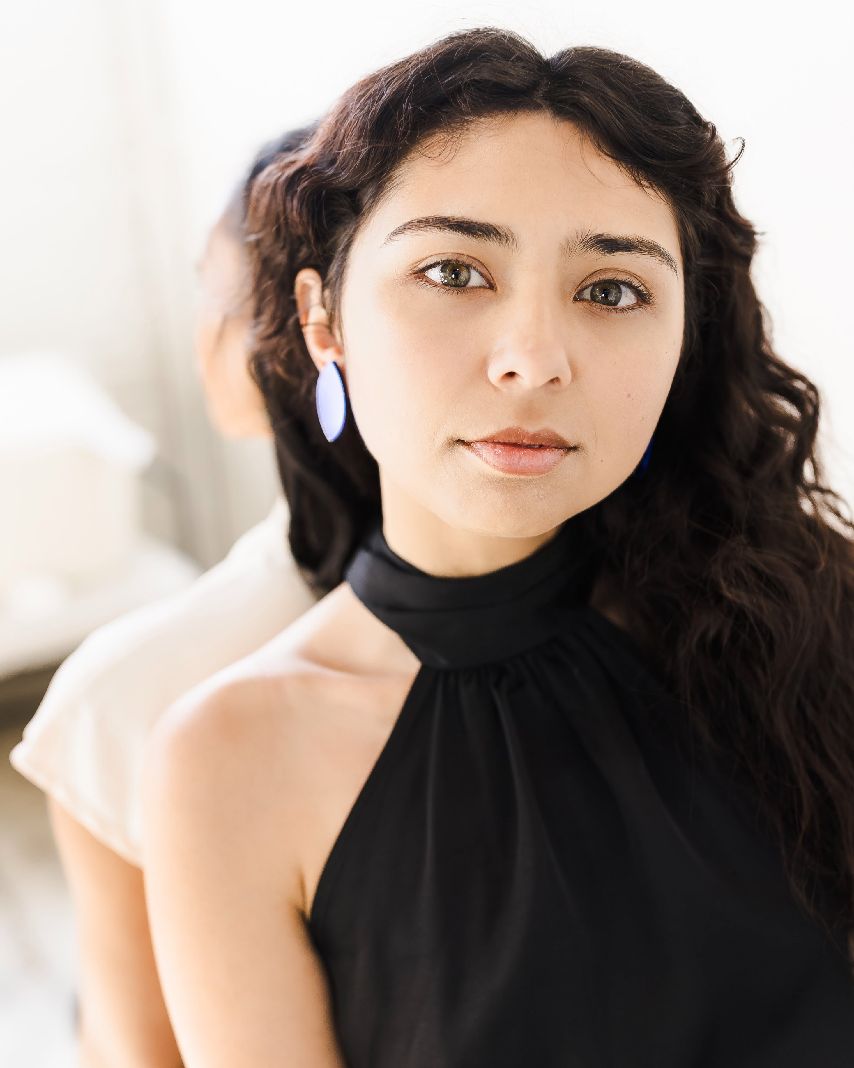 Woman with long, dark, wavy hair in a black halter top and large acrylic earrings looks at the camera. 
