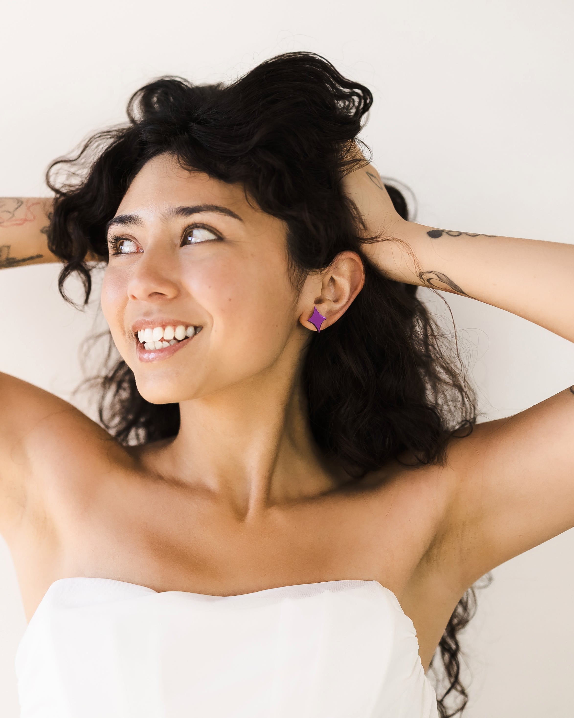 A woman with curly hair wearing a strapless white top, smiling, with her hands behind her head, showcasing her purple earrings and visible tattoos.