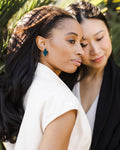 Two women standing close together with a natural background. Woman in forefront wearing geometric earring.
