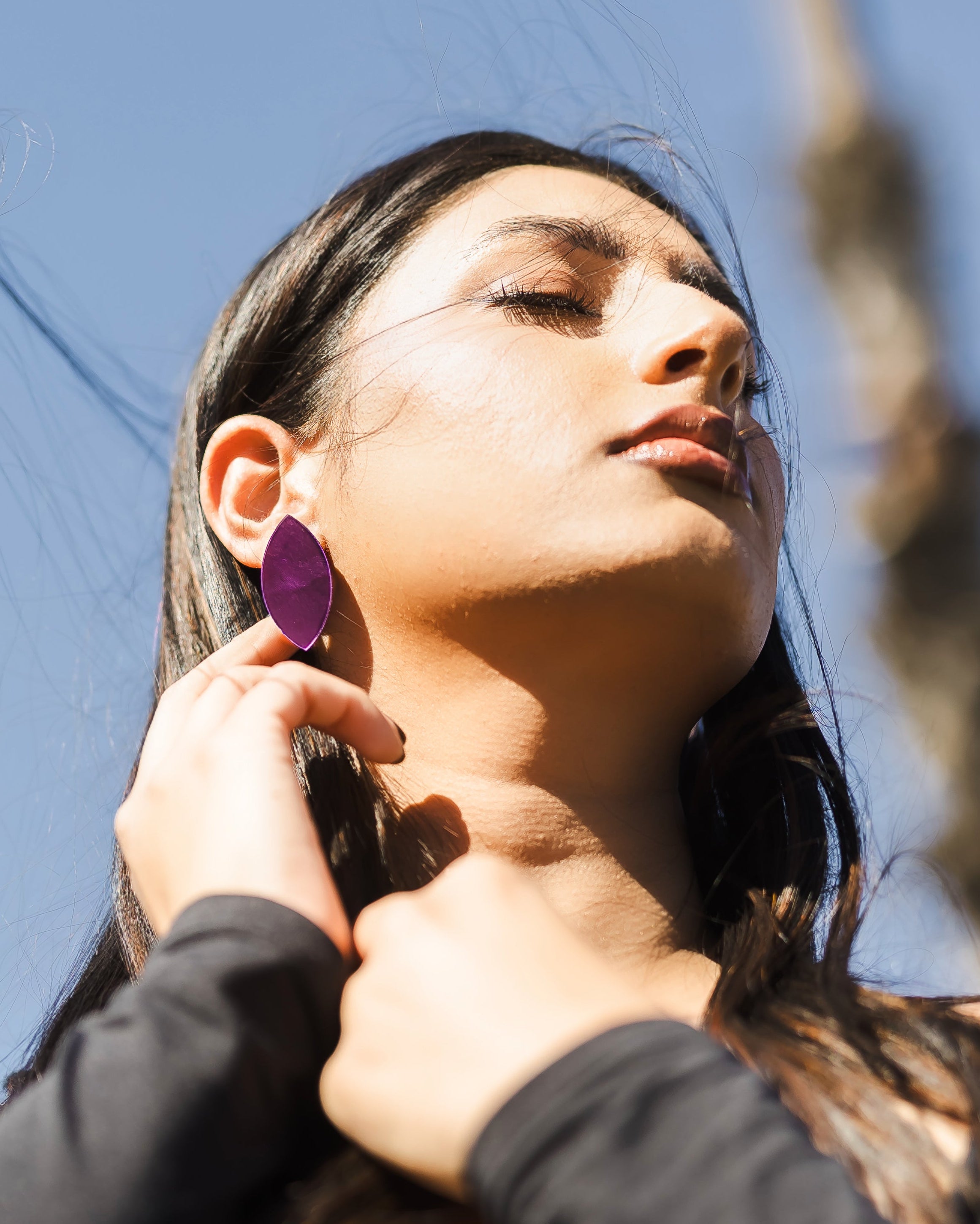 Woman in a black long-sleeve top with windswept hair touches her neck while wearing large purple leaf-shape earrings outside.