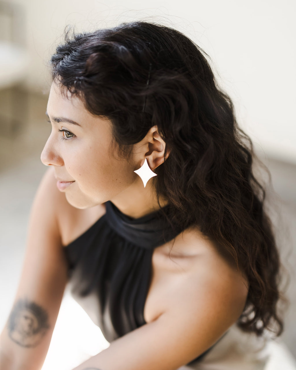 Young woman with long, curly dark hair wearing black sleeveless top and starburst-shaped reflective mirror earrings, with tattoos on inner elbows.