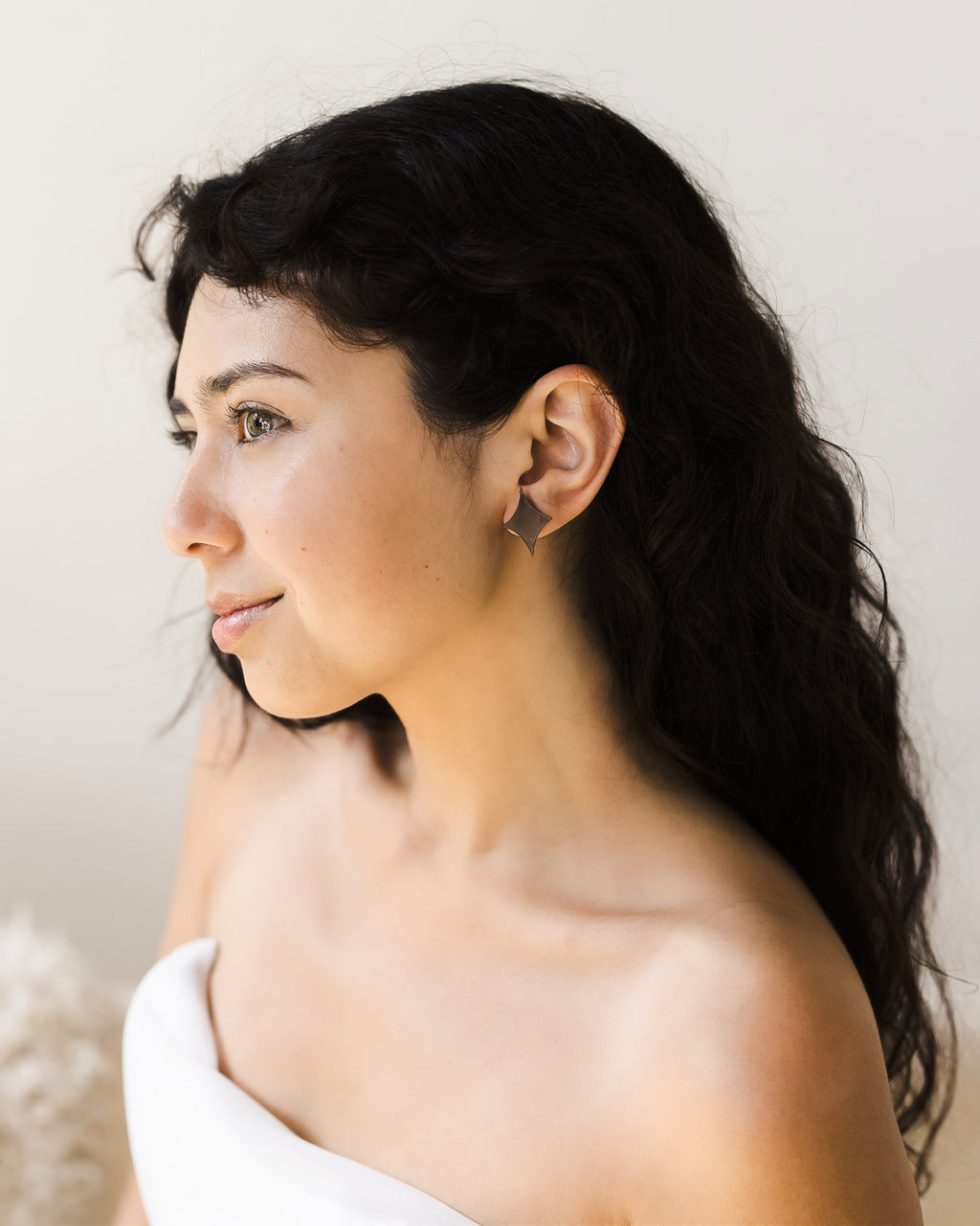 Young woman with long, dark, curly hair wearing a strapless white top, smiling. She wears a small satin gold earring shaped like a 4-point star.
