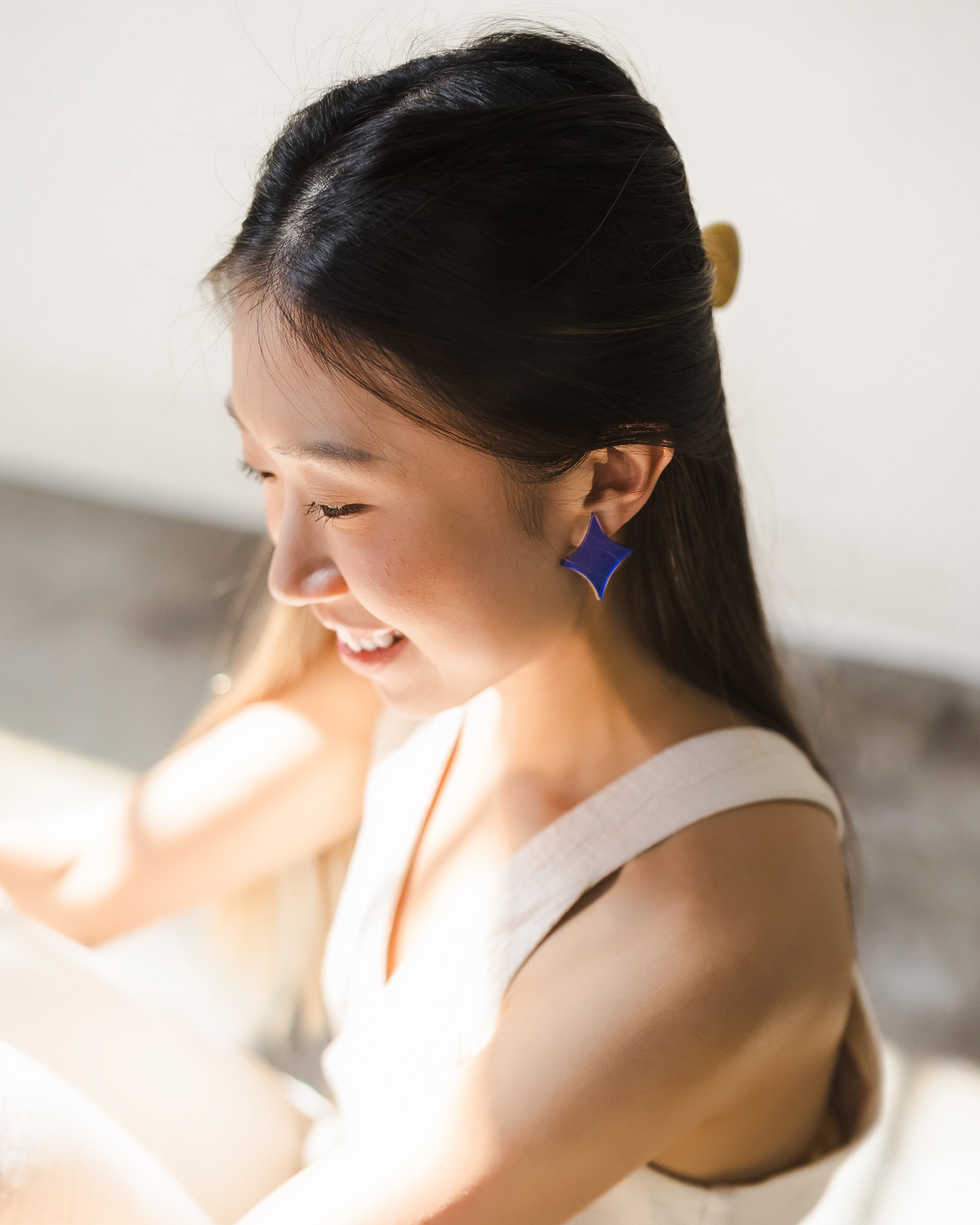 Woman with long, black hair wearing a white top and sapphire- blue 4-point-star-shaped earring on left ear, with soft light falling across her face.