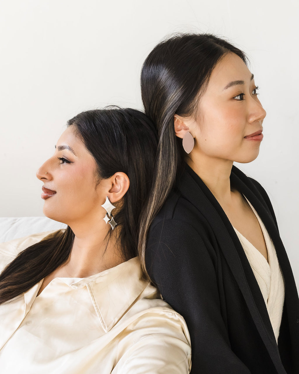 Two women seated back-to-back. One woman is wearing shiny silver statement earrings, while the other woman is wearing neutral matte gold earrings.