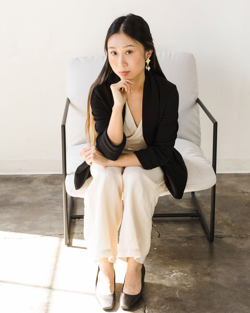 Woman with long hair seated in a light-colored armchair, wearing a black blazer over a beige outfit. She is showcasing silver spark-shape earrings.