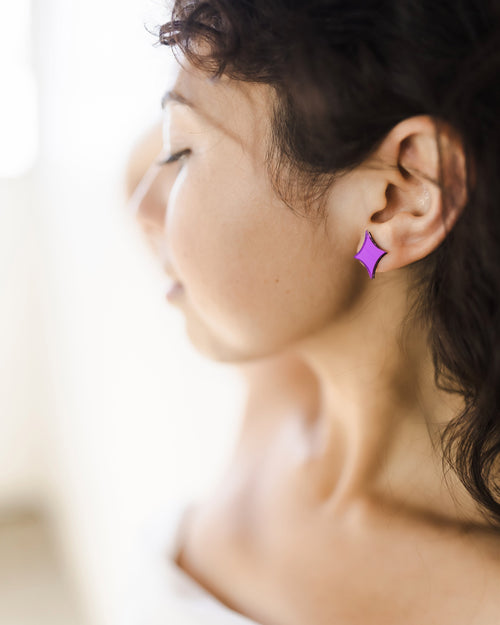 A young woman with curly, dark hair wearing purple acrylic mirror earrings shaped like an AI sparkle icon.