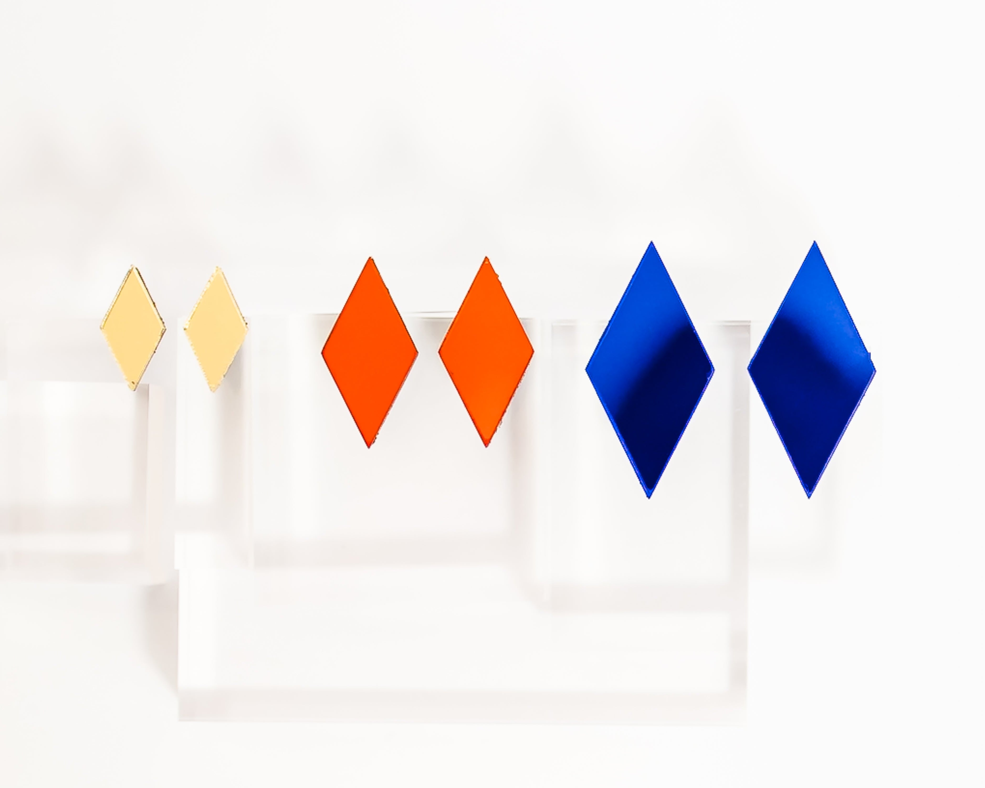 Three pairs of diamond-shaped stud acrylic mirror earrings in golden yellow, red, and blue,  against a white background.