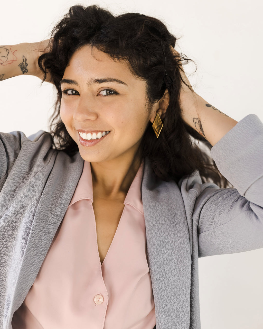 Smiling woman with dark curly hair poses in a light gray blazer over a pale pink button-up blouse, wearing a gold geometric earring.