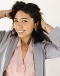 Smiling woman with dark curly hair poses in a light gray blazer over a pale pink button-up blouse, wearing a gold geometric earring.
