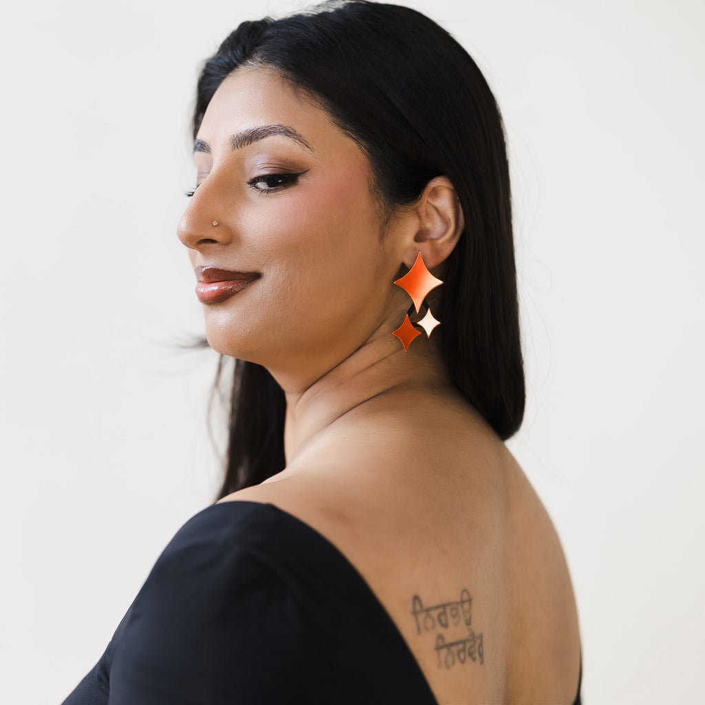 Woman with long black hair side angle, wearing black top. She has a tattoo on her back and is adorned with red four-point-star-shaped earrings.
