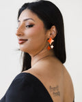 Woman with long black hair side angle, wearing black top. She has a tattoo on her back and is adorned with red four-point-star-shaped earrings.