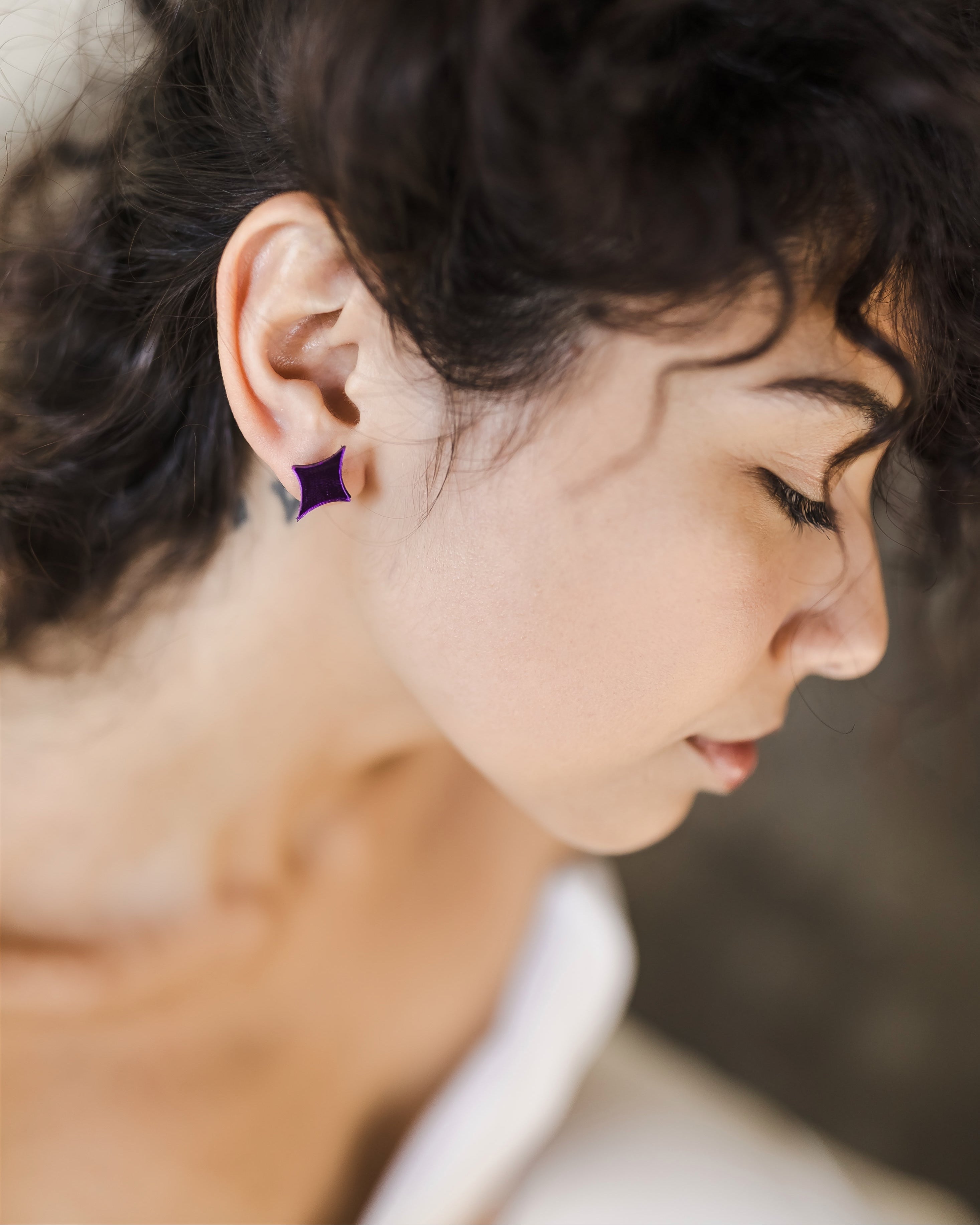 A young woman with curly, dark hair wearing purple acrylic mirror earrings shaped like a spark.