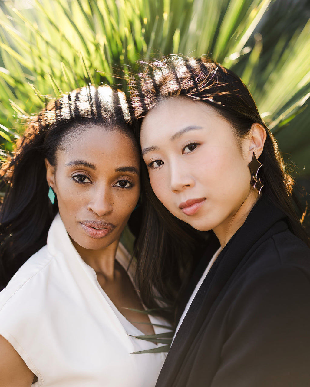 Two women with teal geometric earrings and sparkle reflective earrings against palm trees.